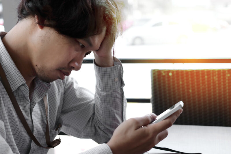 Frustrated Stressed Asian Business Man With Hands On Forehead Looking Mobile Smart Phone In His Hands At Office.