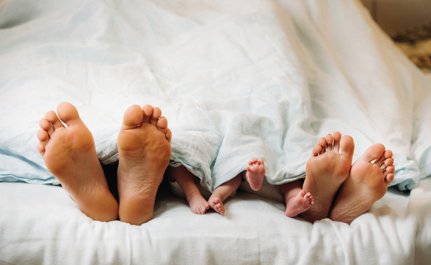 The Feet Of Parents And Young Children Under The Blanket. Mom And Dad With Children Lie Under A Blanket At Home